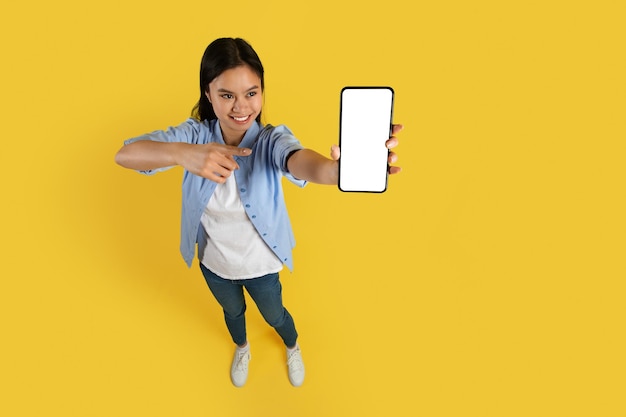 Улыбающаяся забавная молодая китайская студентка в повседневной одежде показывает пальцем на смартфон с пустым экраном