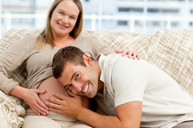 Smiling future dad listening to the belly of his wife sitting on the sofa