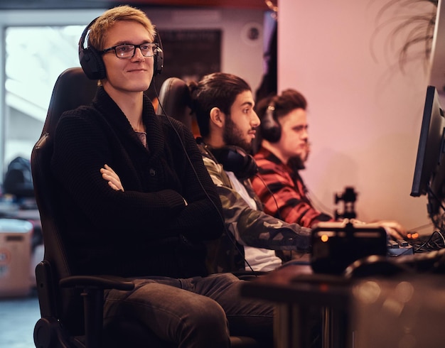 A smiling gamer wearing a sweater and glasses with his arms crossed sitting on a gamer chair in a gaming club or internet cafe.