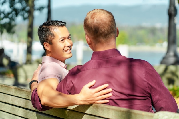 smiling gay men homosexuals sitting on bench hugging looking at each other relaxing outdoors