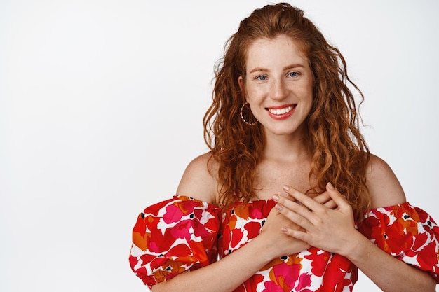 Photo smiling ginger girl in dress holding hands on heart looking pleased being thankful standing against white background