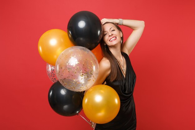 Smiling girl in black dress celebrate putting hand on head hold air balloons isolated on red background. St. Valentine's International Women's Day Happy New Year birthday mockup holiday party concept.