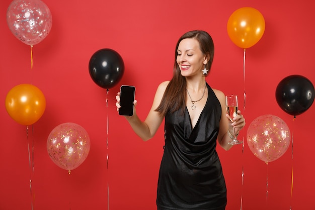 Улыбающаяся девушка в черном платье держит бокал шампанского, глядя на мобильный телефон с пустым черным пустым экраном на ярко-красном фоне воздушных шаров. С Новым годом, концепция праздничной вечеринки макет дня рождения.