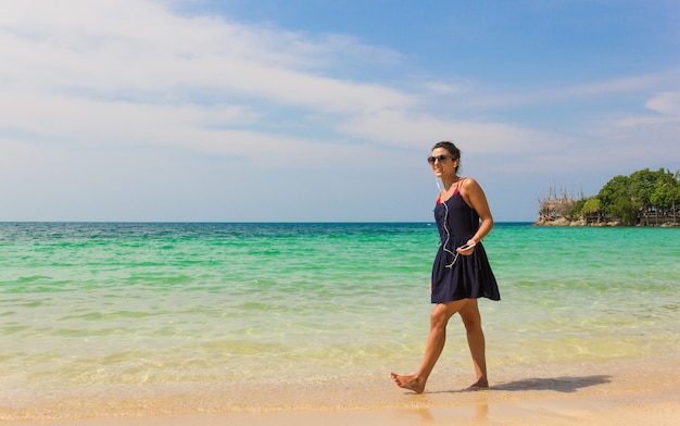 Ragazza sorridente sul vestito blu che ascolta la musica mentre si cammina sulla riva e che tiene un cellulare collegato agli auricolari nell'isola di koh pha ngan, thailandia