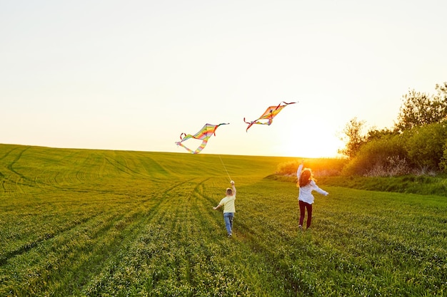 Smiling girl and brother boy running with flying colorful kites on the high grass meadow Happy childhood moments