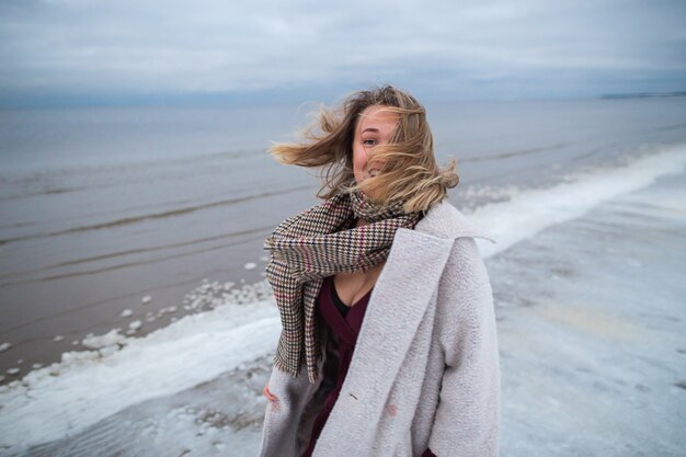 Smiling girl in the burgundy dress and coat on the background\
of the winter sea. portrait of a woman on sea, snow windy weather,\
cold atmospheric image.