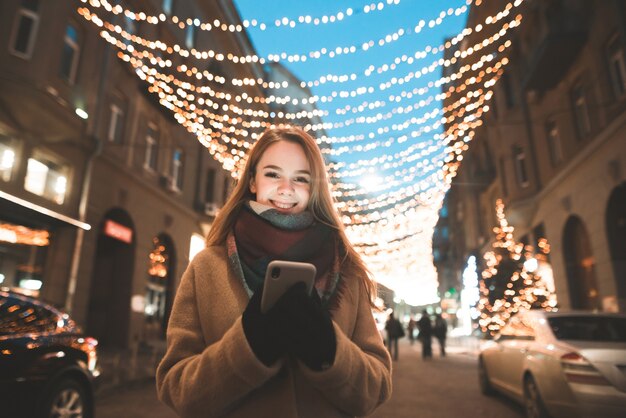 コートで笑顔の女の子と彼の手でスマートフォンは街路灯装飾の背景に立っています。