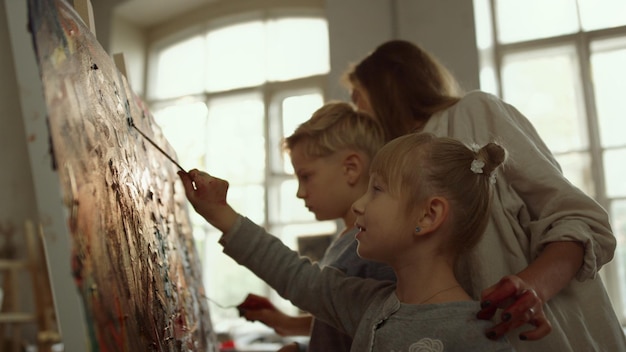 ワーク ショップで絵筆で絵を描く笑顔の女の子 アート スタジオで一緒に時間を過ごす才能のある家族 朝の窓の背景で子供たちを教える創造的な女性アーティスト
