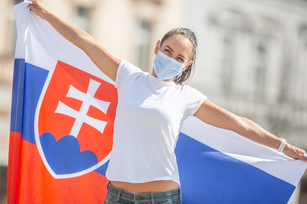 Photo smiling girl in a face mask holds a flag of slovakia behind her on the street.