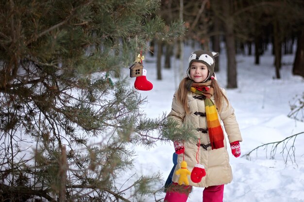 Ragazza sorridente in cappello divertente che decora un albero di pino con un giocattolo artigianale tra la foresta invernale