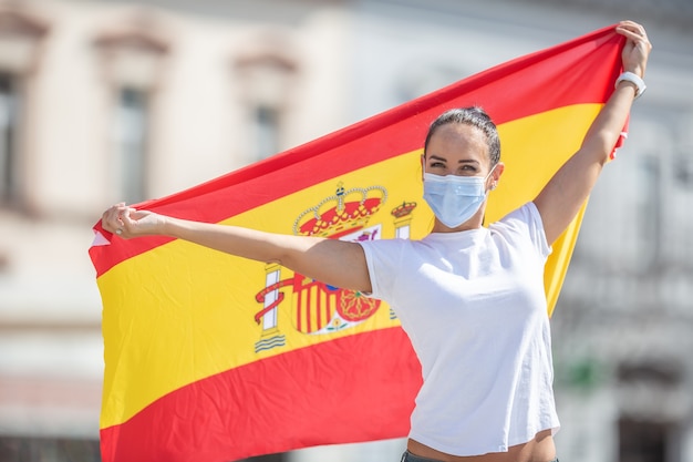 Photo smiling girl holds a spanish flag behind her back wearing a face mask.