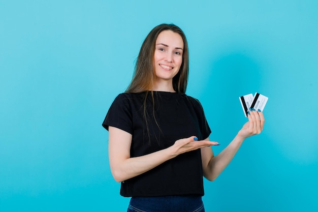 Smiling girl is holding credit cards and showing them with other hand on blue background