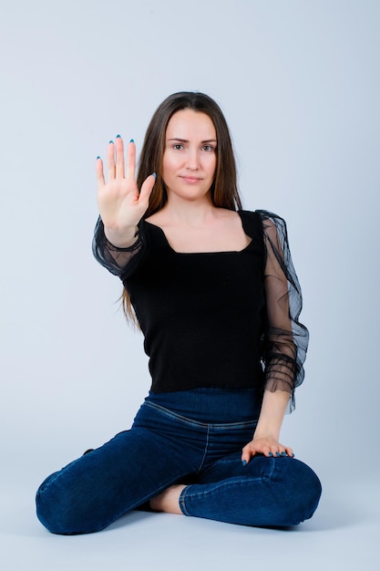 Smiling girl is showing stop gesture by sitting on floor on white background