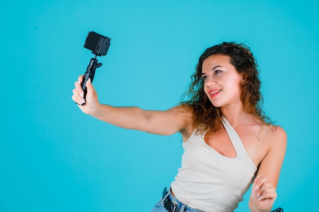 Smiling girl is taking selfie with her mini camera on blue background