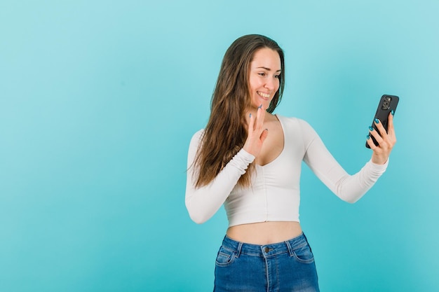 Smiling girl is talking on videocall and showing hi gesture on blue background