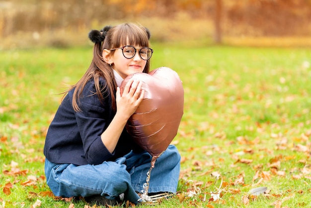 Smiling girl in jeans and jacket holding pink balloon with heart in Autumn park