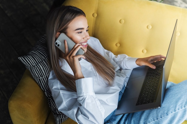 Smiling girl laying on sofa, typing on laptop and talking on phone