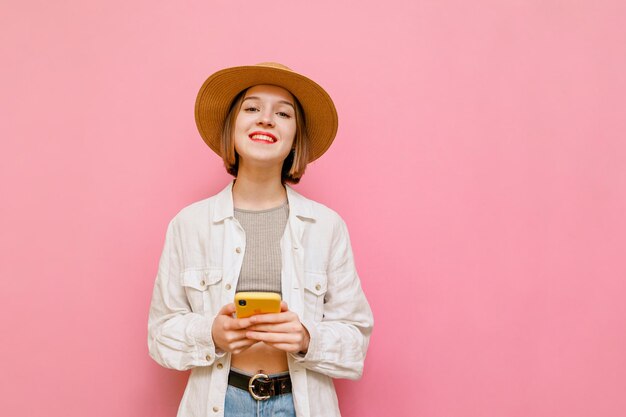 明るい服とピンクの背景の帽子の笑顔の女の子はスマートフォンを保持し、カメラをのぞきます