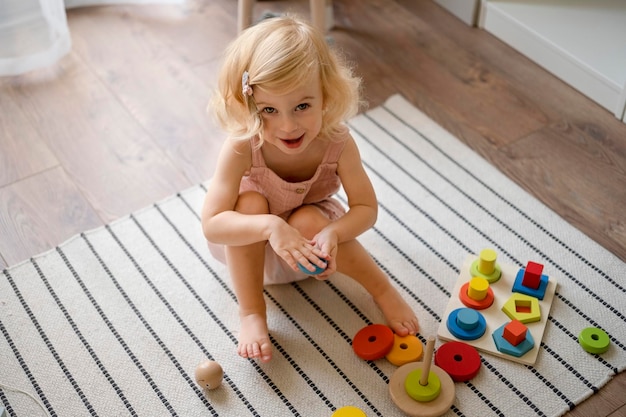 カーペットの上に座ってエコ木のおもちゃで遊んでカメラを見て笑顔の女の子屋内の彼女の部屋で楽しんで幸せな愛らしい幼児キッド