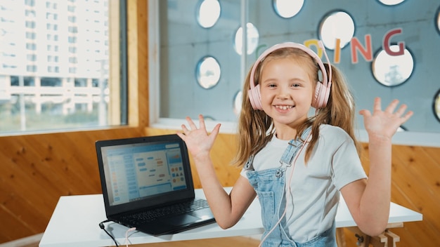 Photo smiling girl looking at camera while waving hand with laptop placed erudition