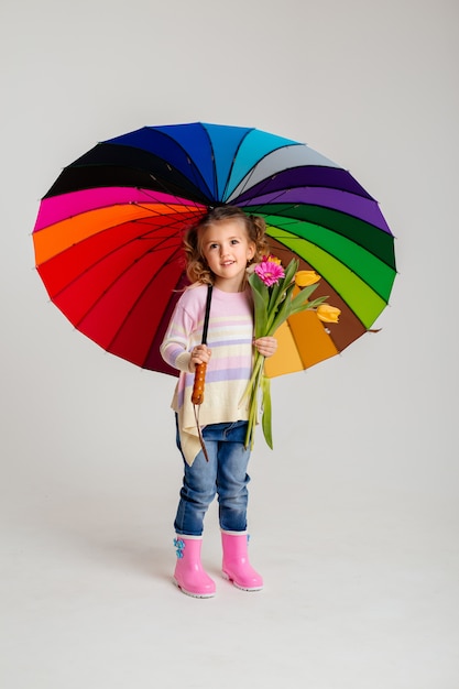Ragazza sorridente nell'abbinamento della camicia rosa e stivali da pioggia che tengono l'ombrello dell'arcobaleno su fondo bianco