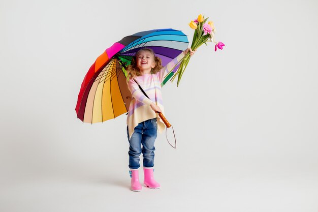 Smiling girl in matching pink shirt and rain boots holding rainbow umbrella on white background