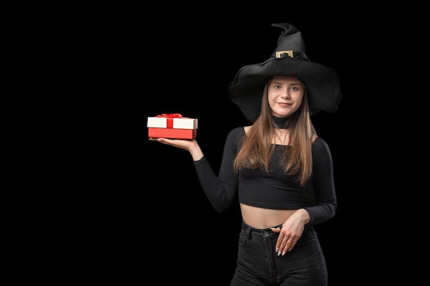 Smiling girl in pointy hat holds gift in her palm Woman with gift Isolation on black background Copy space Halloween