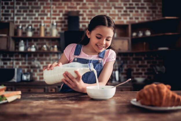 キッチンで朝食のためにコーンフレークのボウルにミルクを注ぐ笑顔の女の子