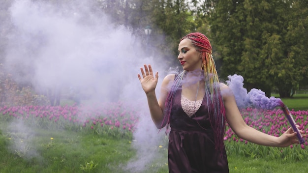 Smiling girl in purple satin dress with long multi-colored braids and eye-catching glitter makeup. Smoke of purple color covers the girl in the spring park