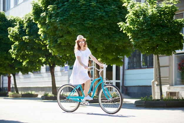 青いビンテージバイクに乗って、緑の木々や花壇でカメラを探している笑顔の女の子