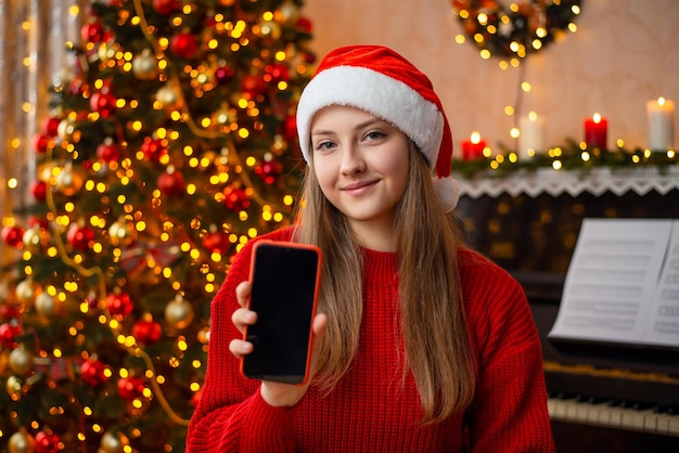 Улыбающаяся девушка в шляпе Санты и красном свитере показывает экран телефона на камеру Портрет молодой девушки с телефоном