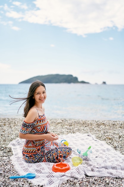 Smiling girl sits on a bedspread on a pebble beach with childrens toys