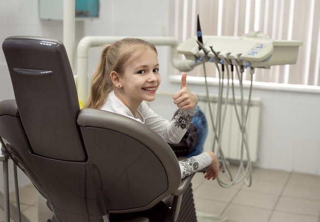 A smiling girl sits in a dental chair and gives a thumbsup