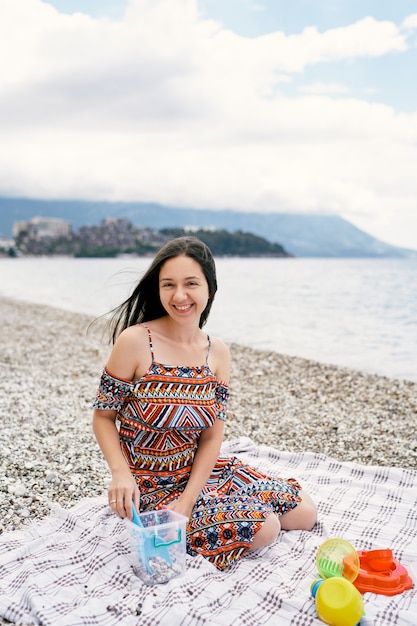 Smiling girl sitting on a blanket on a pebble beach with toys