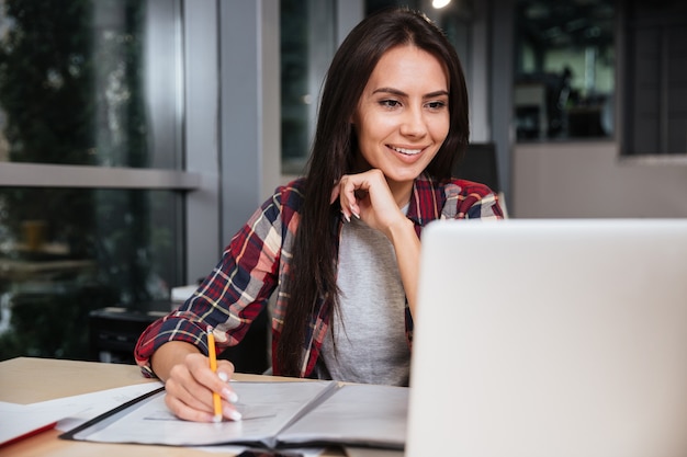 Smiling Girl sitting by the table with documents and laptop.