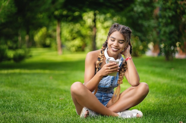 Ragazza sorridente che si siede sull'erba nel parco e scrive un messaggio sul telefono