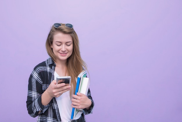 Photo smiling girl stands against a purple wall with books in her hands and uses a smartphone.