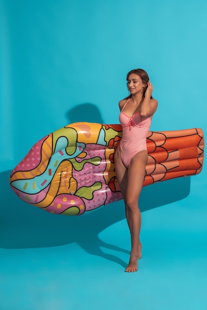 Smiling girl in swimsuit carrying inflatable mattress on blue wall