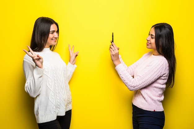 Smiling girl taking a photo of her girlfriend isolated over yellow wall