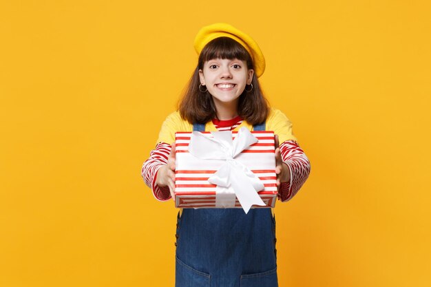 Smiling girl teenager in french beret, denim sundress holding red striped present box with gift ribbon isolated on yellow wall background. People sincere emotions, lifestyle birthday holiday concept.