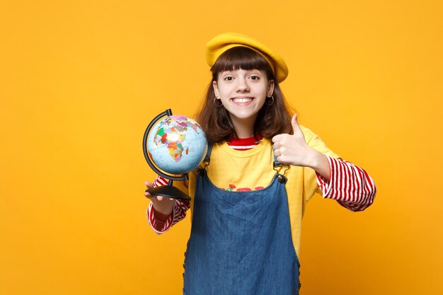 Smiling girl teenager in french beret and denim sundress showing thumb up, holding Earth world globe isolated on yellow wall background. People sincere emotions, lifestyle concept. Mock up copy space.