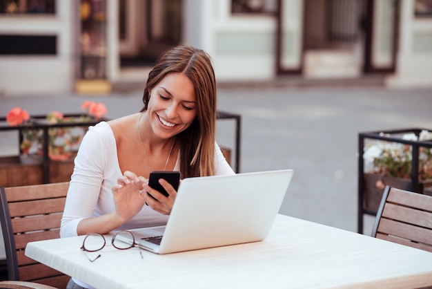 Улыбающаяся девушка с помощью телефона и ноутбука в кафе на открытом воздухе.