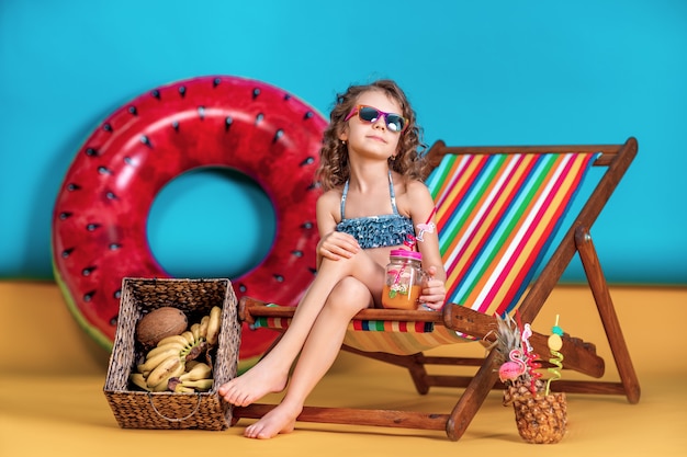 Smiling girl wearing swimsuit and sunglasses holding jar with juice or cocktail with multicolored straws sitting in rainbow deck chair