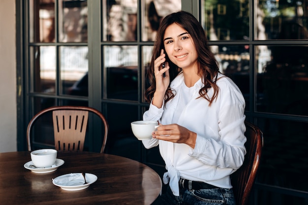 Smiling girl in white shirt holds cup and talks in cafe