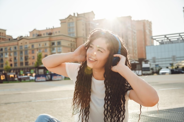 ヘッドフォンでドレッドヘアを持つ笑顔の女の子が公園の床に座っています ヘッドフォンで音楽を楽しんでリラックスする幸せな若い女性 世代 z をコピーするためのスペース