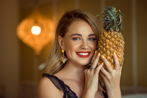 Smiling girl with evening make up wearing accessories posing and holding pineapple, close up.