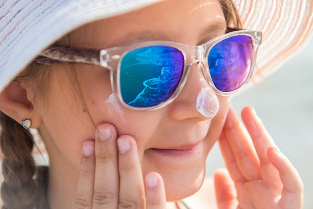 帽子と微笑んでいる女の子がビーチで保護フェイスクリームを塗ります