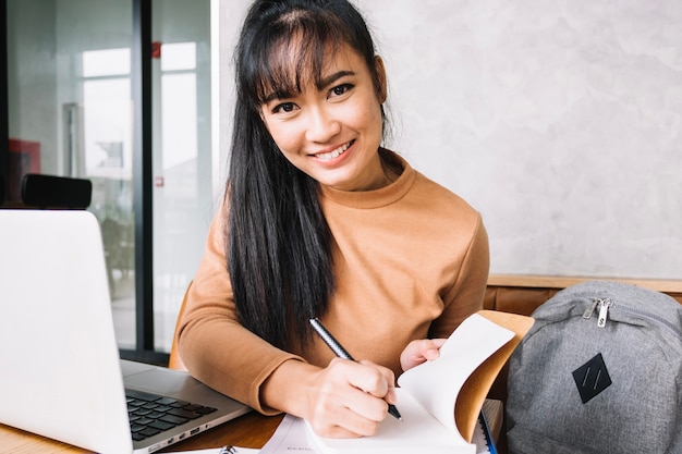 Smiling girl with laptop writing in notebook