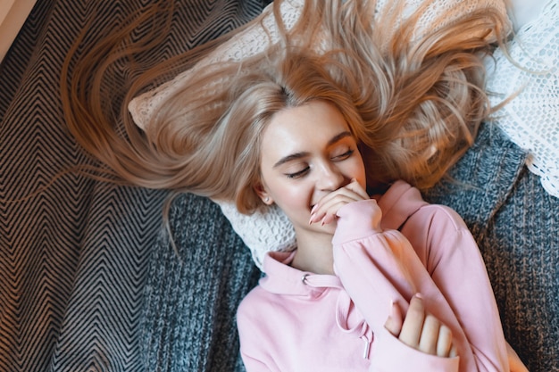 Smiling girl, with long blond hair, enjoying happiness while lying on bed at home