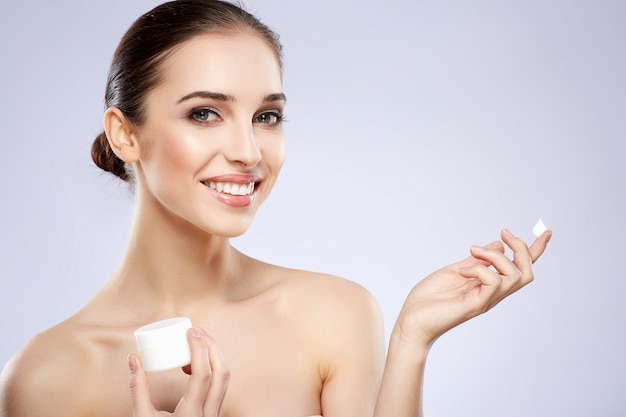 Smiling girl with nude make up posing at grey studio background, beauty photo concept, perfect skin, holding product, portrait.